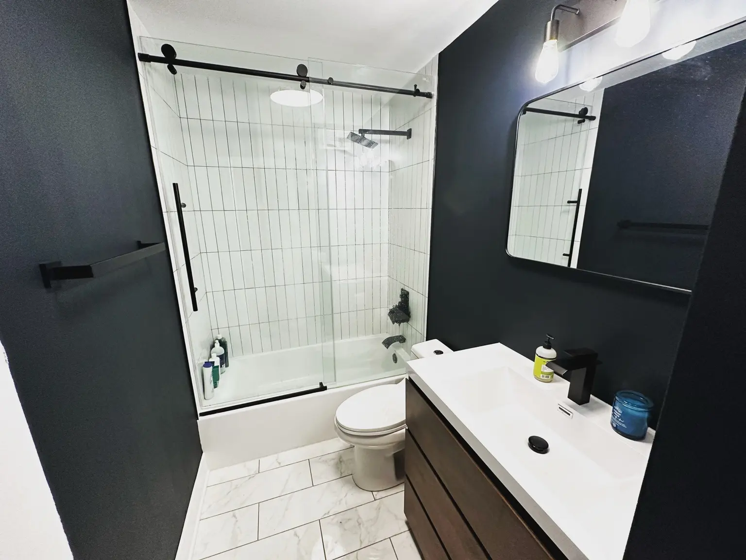 Completed bathroom remodel with black walls, white vertical tile, a frameless sliding glass shower, matte black fixtures and a wood vanity