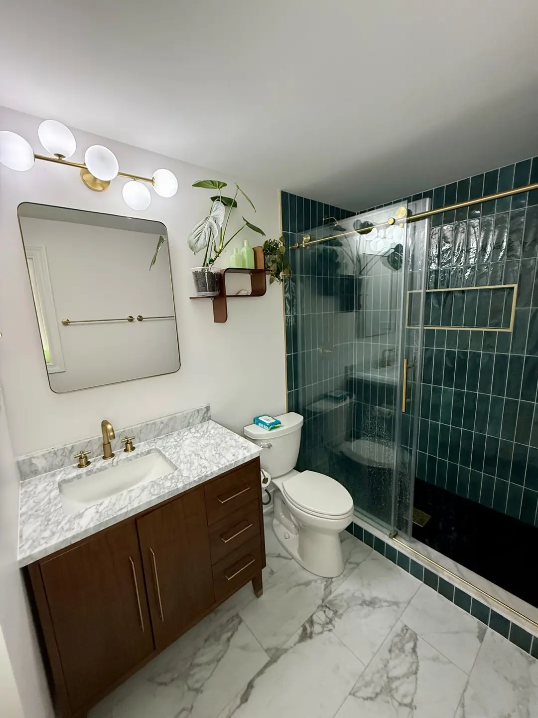 Completed bathroom remodel with deep green tile, a frameless glass shower, brass fixtures, marble-look flooring and a walnut vanity