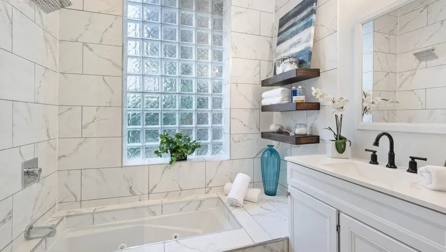 Completed bathroom remodel with marble-look tile, glass-block window, jetted tub, floating wood shelves and white vanity