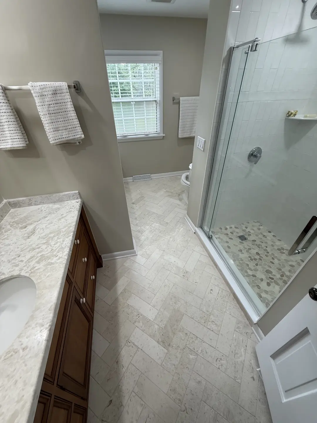 Completed bathroom remodel with a frameless glass shower, white subway tile, pebble shower floor, travertine herringbone floor and wood vanity
