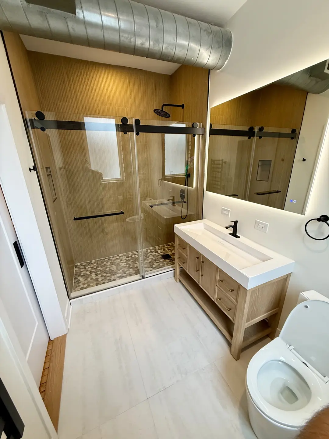 Completed bathroom remodel with a warm wood shower surround, matte black sliding glass door, pebble mosaic shower floor and light oak vanity