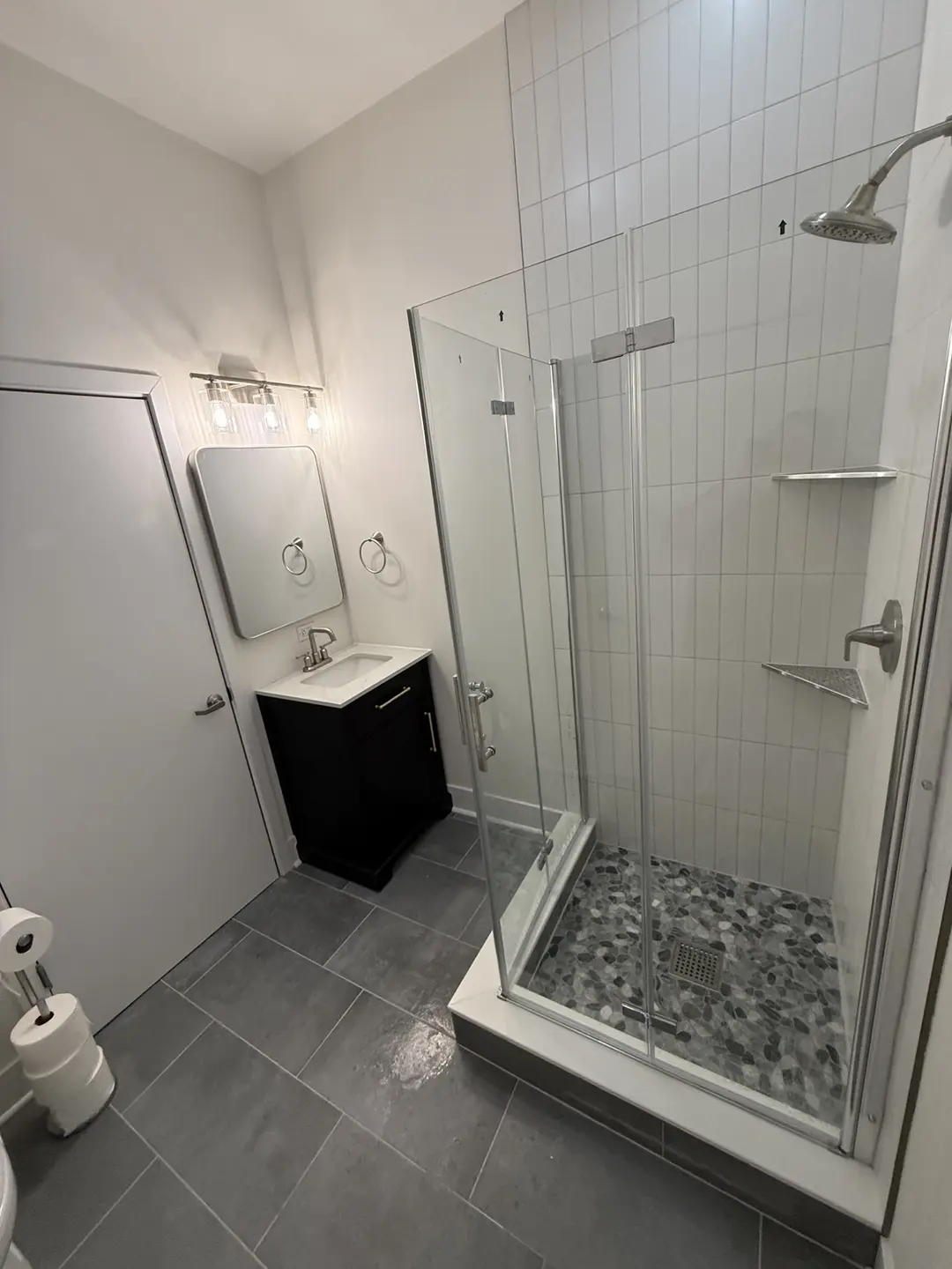 Completed bathroom remodel with a frameless glass shower, vertical white tile, pebble shower flooring, a black vanity and charcoal porcelain floor tile